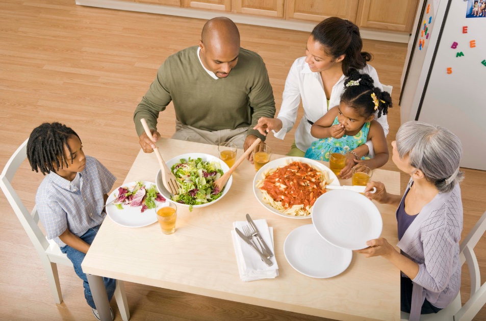 A family of 5 sitting around the dinner table. They are enjoying pasta and the father is serving salad. 